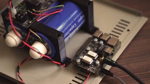 Raspberry Pi mit aufgestecktem Transport-HAT auf einer Gehaeusegrundplatte, links die Supercap-Stromversorgung mit blauen Ultrakondensatoren, verbunden ueber farbige Litzen